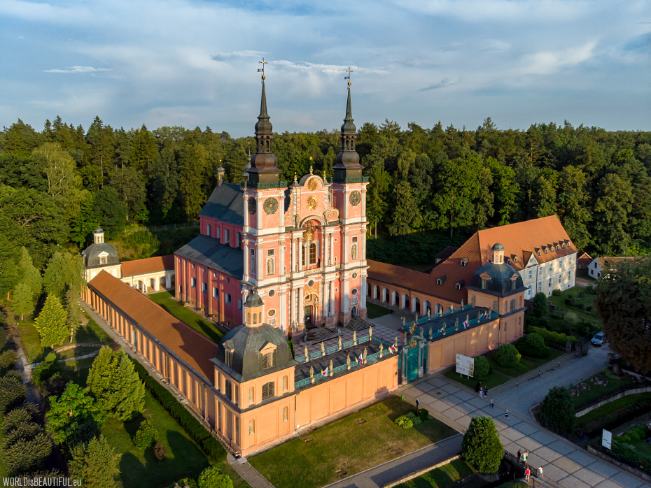 Ujęcie z drona przedstawiające barokowy kompleks klasztorny Wniebowzięcia Najświętszej Maryi Panny i św. Józefa w Krzeszowie, w otoczeniu gęstych lasów. Centralnym elementem jest główny kościół o różowej fasadzie z dwoma strzelistymi, czarnymi wieżami z hełmami. Kompleks otoczony jest murem z brzoskwiniowej cegły i długimi, brązowymi skrzydłami klasztornymi. W prawym dolnym rogu widoczny jest fragment ogrodu i aleja spacerowa. Niebo jest częściowo zachmurzone, a słońce rzuca ciepłe, wieczorne światło.